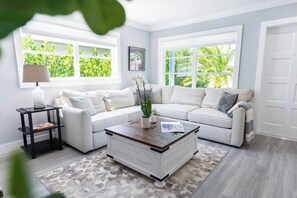 A living room with a white sofa set, a coffee table, and a view of greenery through large windows. - 778 De La Sol • Southern Exposure Heated Pool Home (Naples)