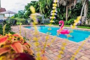 A swimming pool with a pink flamingo float, surrounded by a brick patio and lush greenery. - 563 Park Place | Bougain"Villa" | Mins to Beaches (Naples)