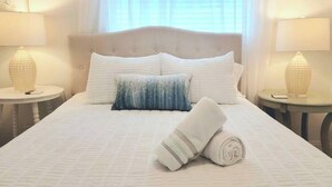Una cama bien tendida con una colcha blanca, dos almohadas y una toalla enrollada. Hay dos mesitas de noche con lámparas.