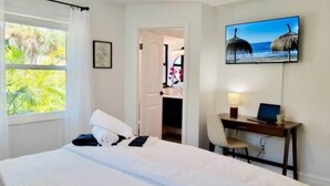 Hotellihuone, jossa on sänky, työpöytä kannettavalla tietokoneella, televisio ja ikkuna, josta avautuu näkymä palmuille.