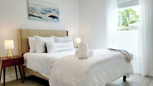 Una cama bien hecha con ropa blanca, una lámpara de noche y una obra de arte colgada en la pared.