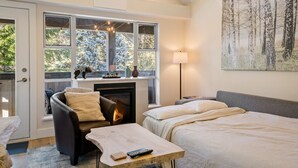 Une chambre confortable avec une cheminée, une grande fenêtre donnant sur les arbres, un lit, une chaise et une table de chevet.