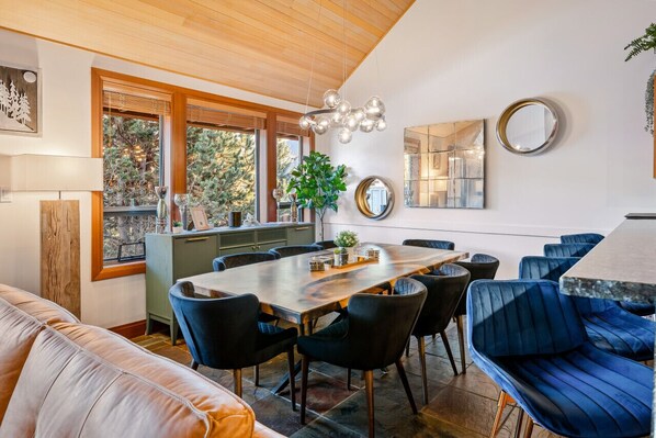 Un espace repas moderne avec une table en bois, des chaises recouvertes d’un tissu bleu et une grande fenêtre donnant sur la forêt.
