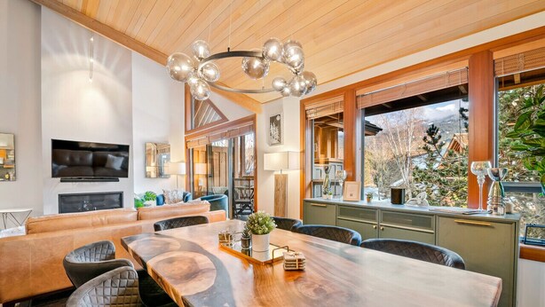Ein modernes Wohnzimmer mit großem Holzesstisch, bequemen Sitzgelegenheiten, einem Kamin und einem großen Fenster mit Blick ins Freie.