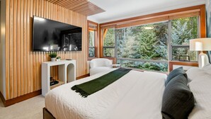 Ein modernes Schlafzimmer mit einem großen Bett, einem an der Wand montierten Fernseher, einem Fenster mit Blick auf Bäume und einem kleinen weißen Konsolentisch.