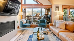 Ein modernes Wohnzimmer mit großem Fernseher, bequemen Sitzmöglichkeiten, einem Kamin und Blick auf die Berge.