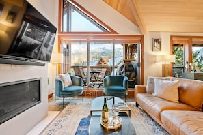 A modern living room with a large TV, two blue armchairs, a brown sofa, and a glass coffee table with a bottle and glasses. - Whistler Village North: 4BR+3.5BA Private Hot Tub (Whistler)