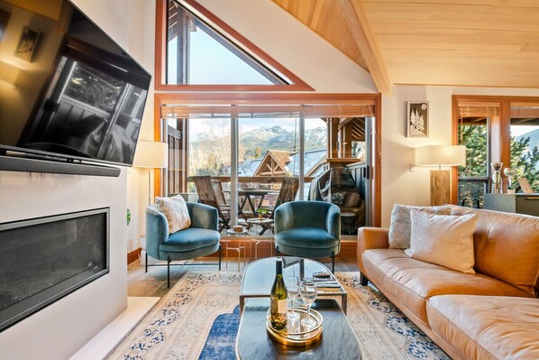 A modern living room with a large TV, two blue armchairs, a brown sofa, and a glass coffee table with a bottle and glasses.