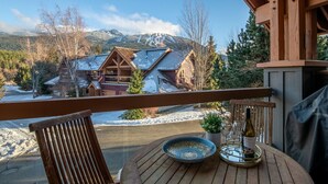 Eine Holzdiele mit Esstisch, mit Blick auf eine Berghütte mit schneebedecktem Dach und blauem Himmel.