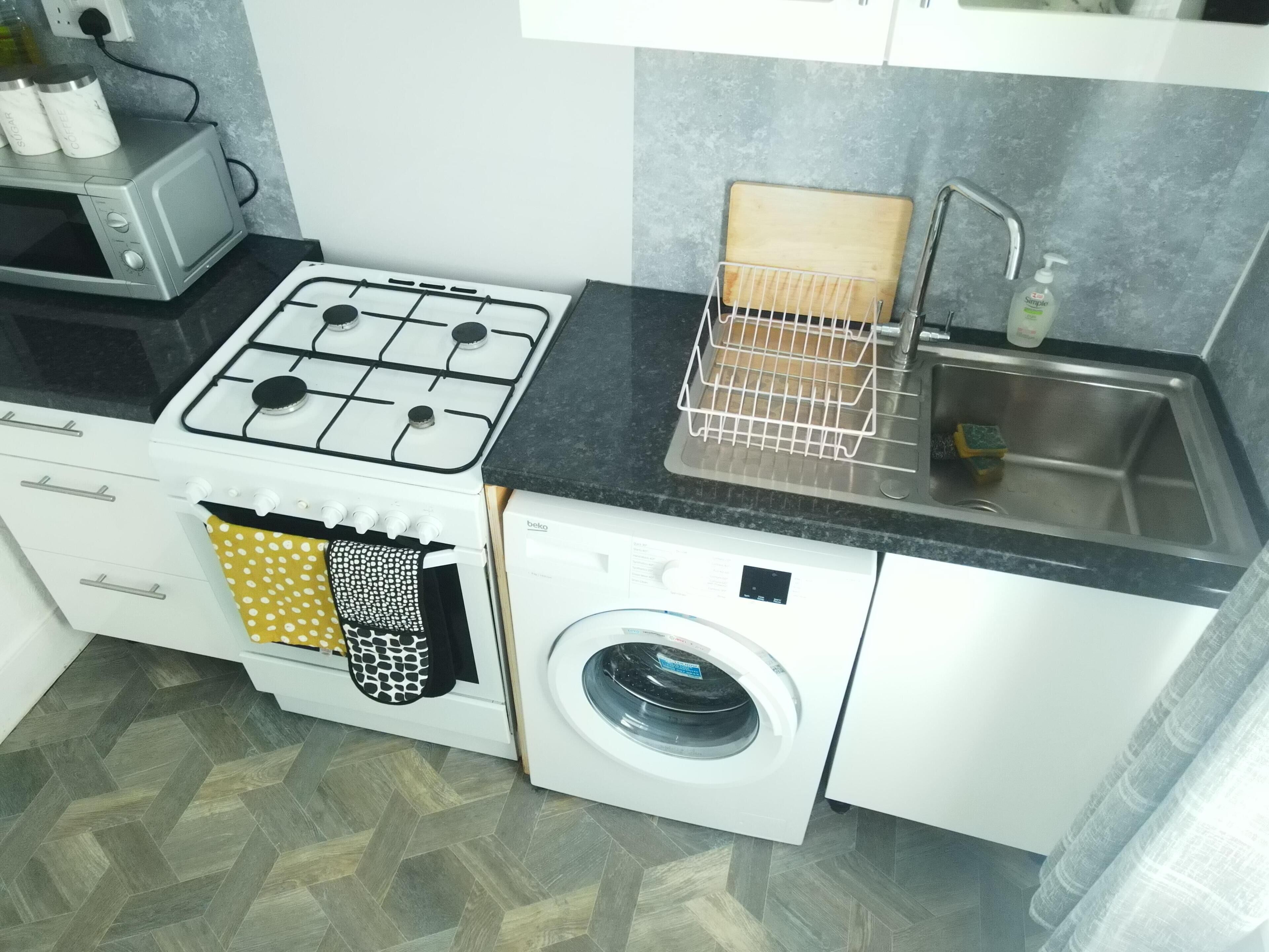 A compact kitchen with a gas stove, a washing machine, a sink, and a drying rack.