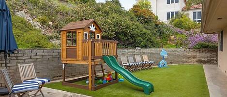 A backyard with a wooden playhouse, a green slide, artificial grass, and a brick wall.