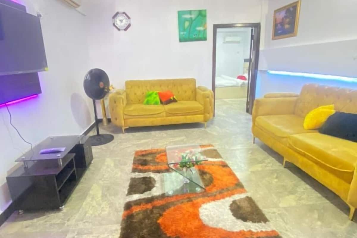 Una sala de estar con dos sofás amarillos, una mesa de centro de vidrio y una alfombra estampada.