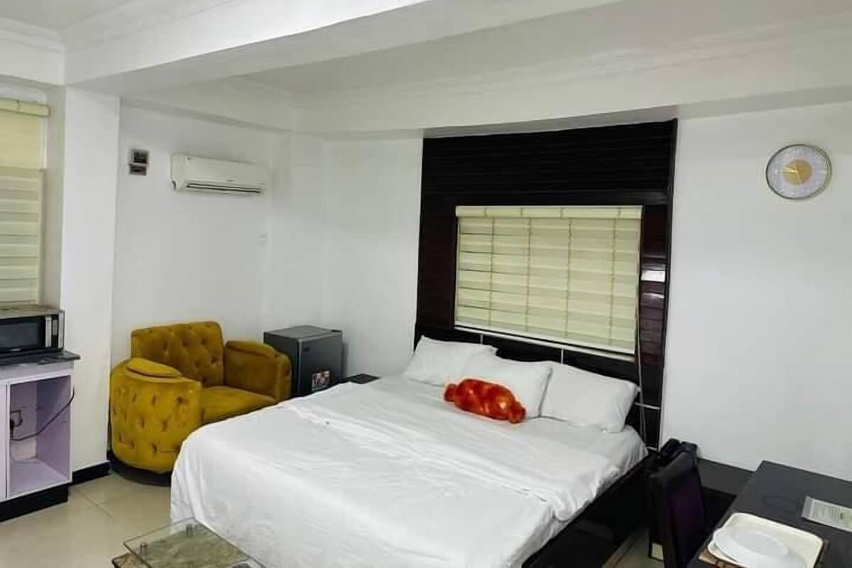Habitación de hotel con cama, un sillón amarillo, un escritorio pequeño con una computadora y una ventana con persianas.