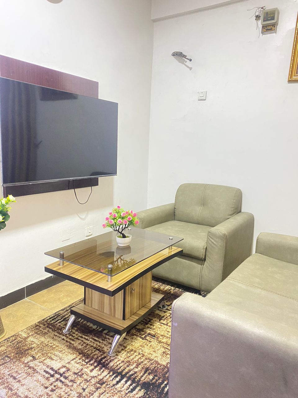 A living room with a flat-screen TV mounted on the wall, a glass-top coffee table, a beige sofa, and a patterned rug.