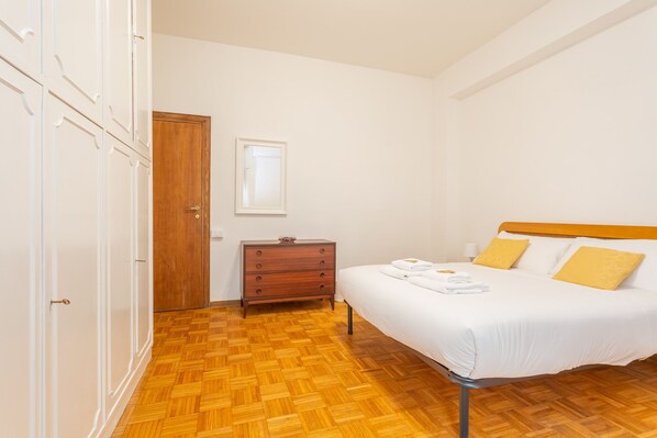 Une chambre avec un plancher en bois, un lit avec une literie blanche et des oreillers jaunes, une commode en bois et une porte en bois.