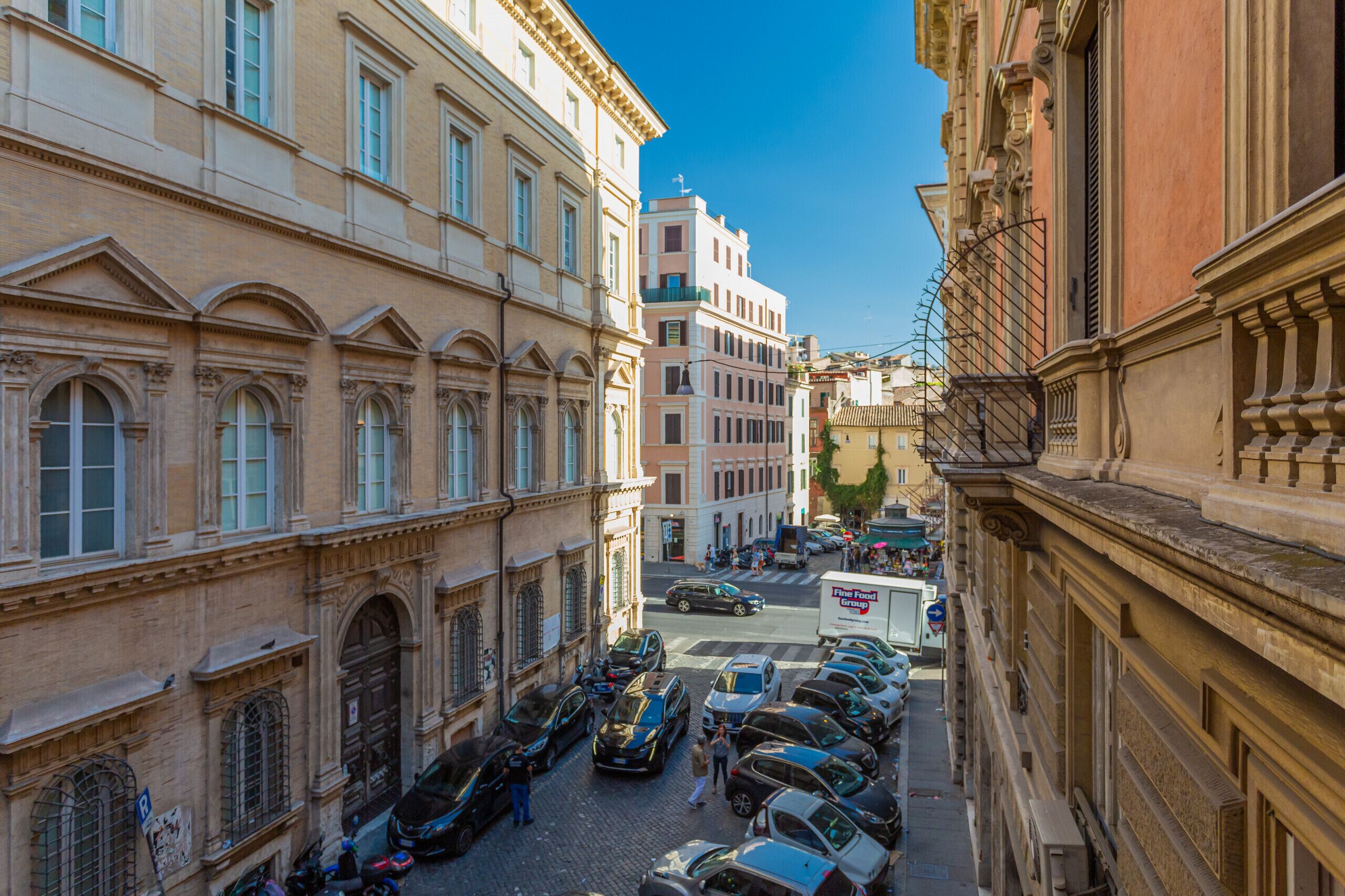 A narrow street lined with historic buildings, parked cars, and a visible Fine Food Group truck.