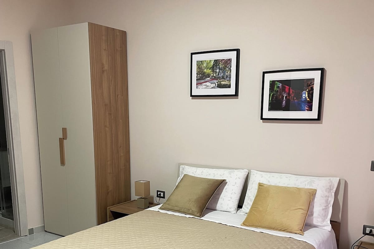 A neatly made bed with a quilted cover, a wooden wardrobe, and two framed pictures on the wall.
