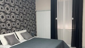 A neatly made bed with a gray quilt, a patterned wall, and a window with sheer curtains.