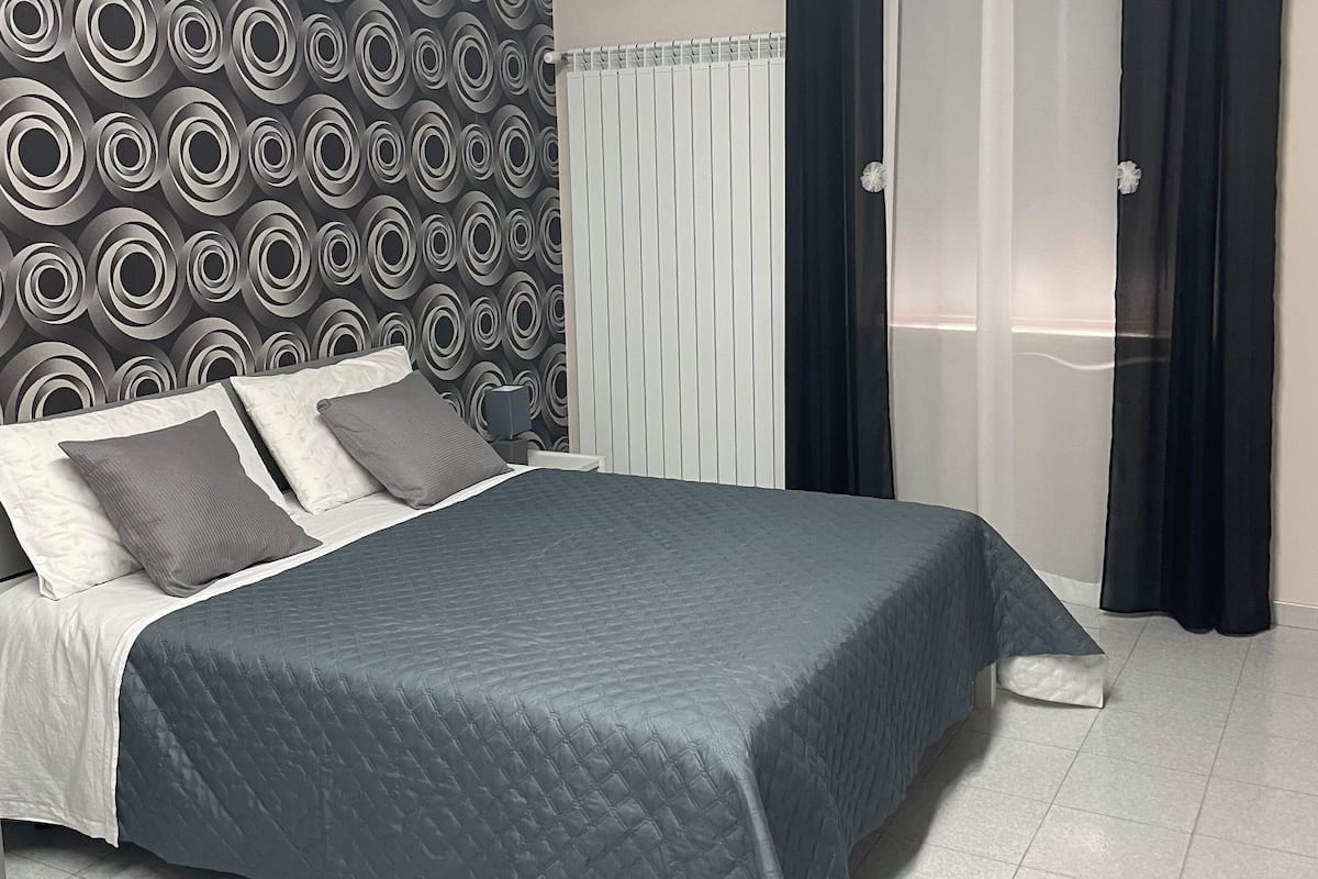 A neatly made bed with a gray quilt, a patterned wall, and a window with sheer curtains.