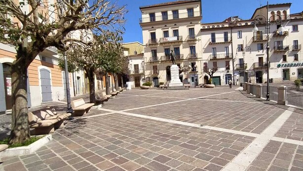 A paved square with benches and trees, surrounded by multi-story buildings with balconies and storefronts.