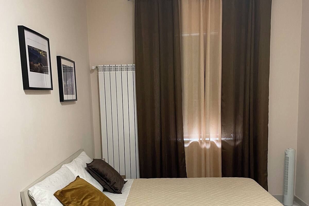 A neatly arranged bedroom with a bed, bedside tables, a radiator, and curtains.