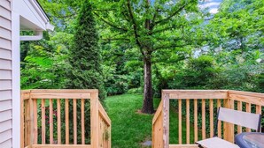 Une terrasse en bois avec un banc et un barbecue, entourée d’une végétation luxuriante et d’arbres.