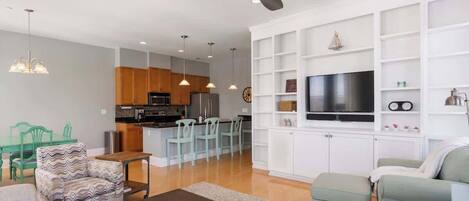 A modern living room with a sofa, armchair, and coffee table, a kitchen area with wooden cabinets and a dining set, and a TV mounted on a white wall.