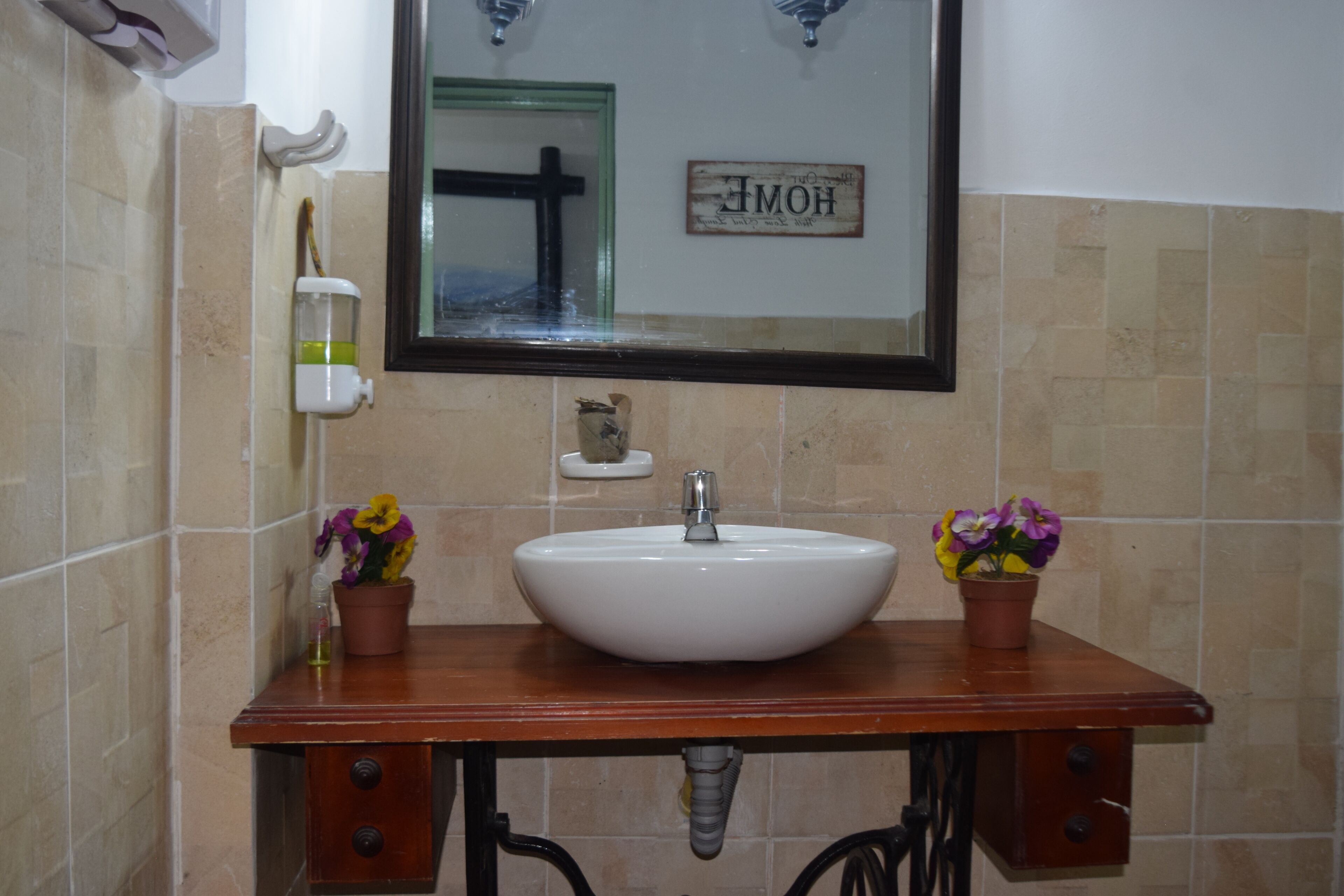 Une salle de bain avec un lavabo blanc, deux pots de fleurs, un miroir et une pancarte indiquant « MAISON ».