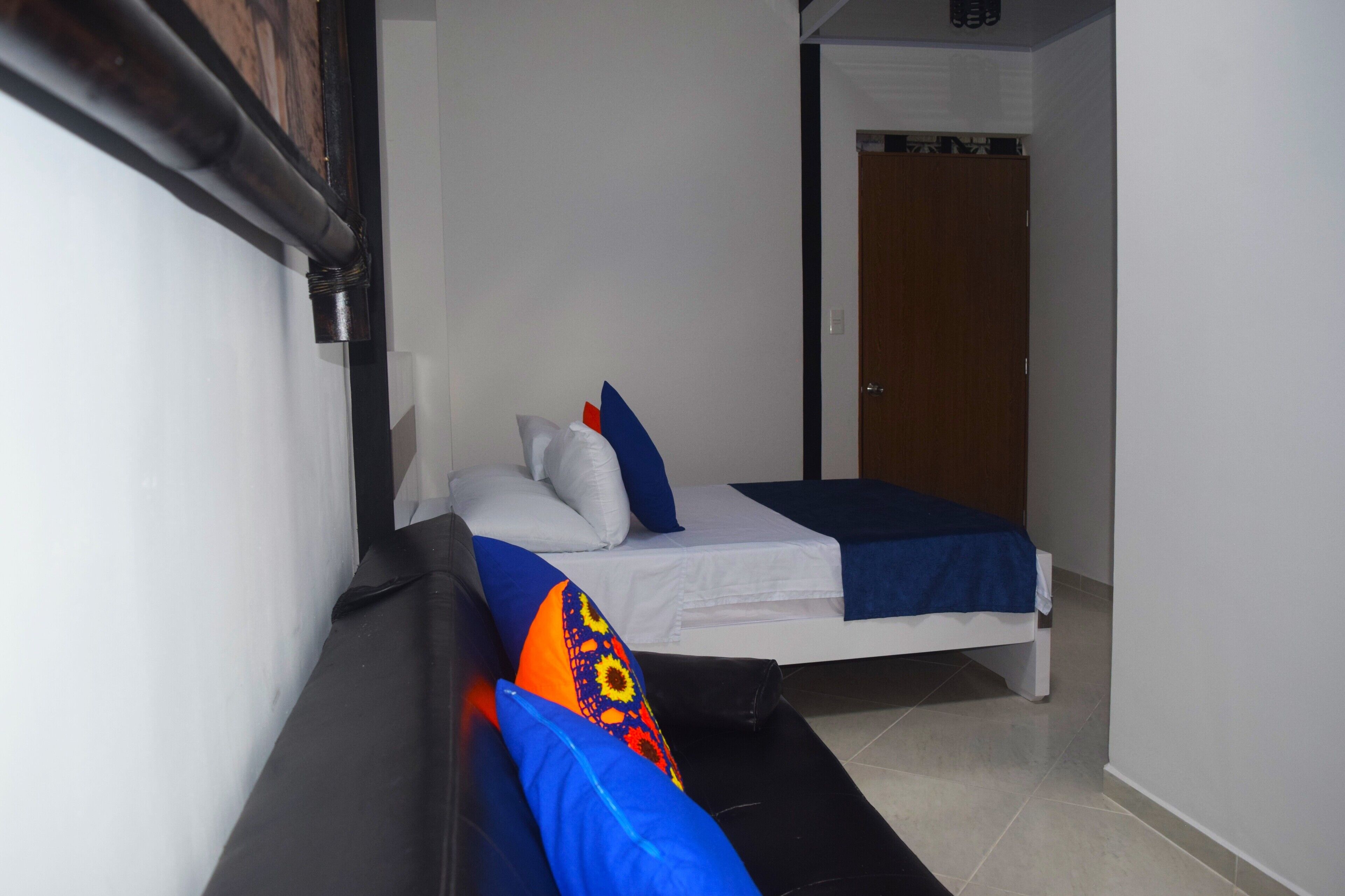 Quarto de hotel pequeno e estreito, com uma cama de solteiro, um banco e uma porta que dá para outro cômodo.