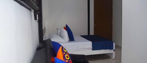 Quarto de hotel pequeno e estreito, com uma cama de solteiro, um banco e uma porta que dá para outro cômodo.