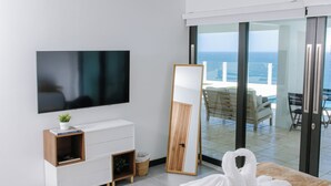 Une chambre d’hôtel moderne dotée d’un grand lit, d’un téléviseur à écran plat, d’une table de chevet agrémentée d’une plante, d’une table d’appoint en bois et offrant une vue sur l’océan grâce à une porte-fenêtre coulissante.