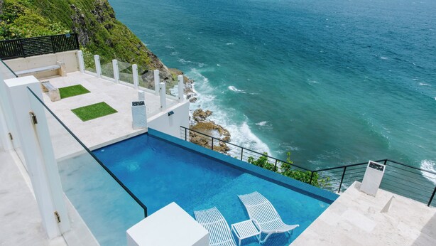 Une piscine bordée de chaises longues blanches, avec vue sur l’océan et une falaise rocheuse en arrière-plan.