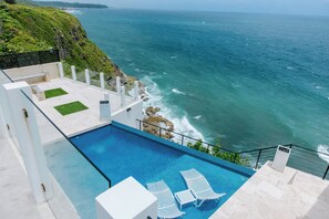 A pool with white lounge chairs, a view of the ocean, and a rocky cliff.