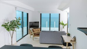 Un salon moderne avec un canapé, une télévision et une table à manger, de grandes fenêtres donnant sur la mer et des plantes en pot.
