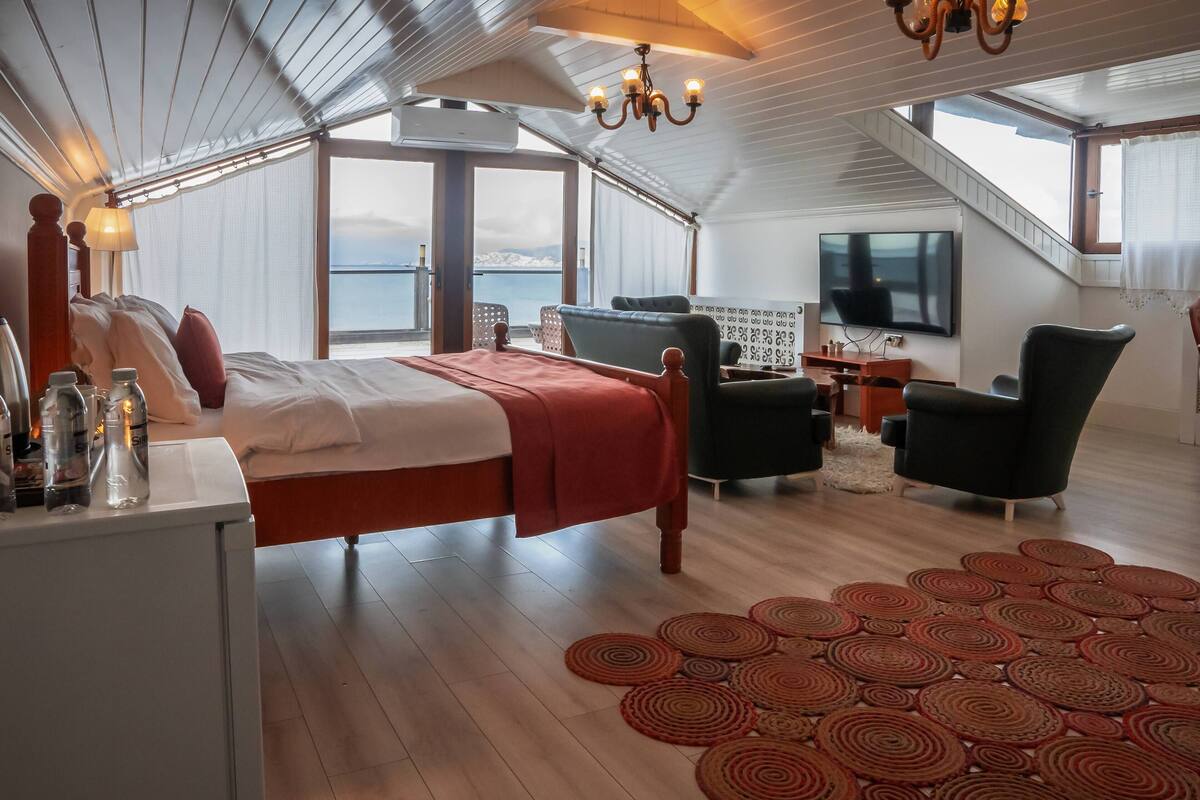A bedroom with a bed, a desk with bottles, a TV, and a view of the sea.