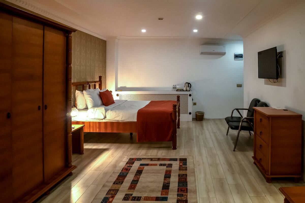 A bedroom with a bed, a wooden wardrobe, a desk, a chair, and a television on the wall.