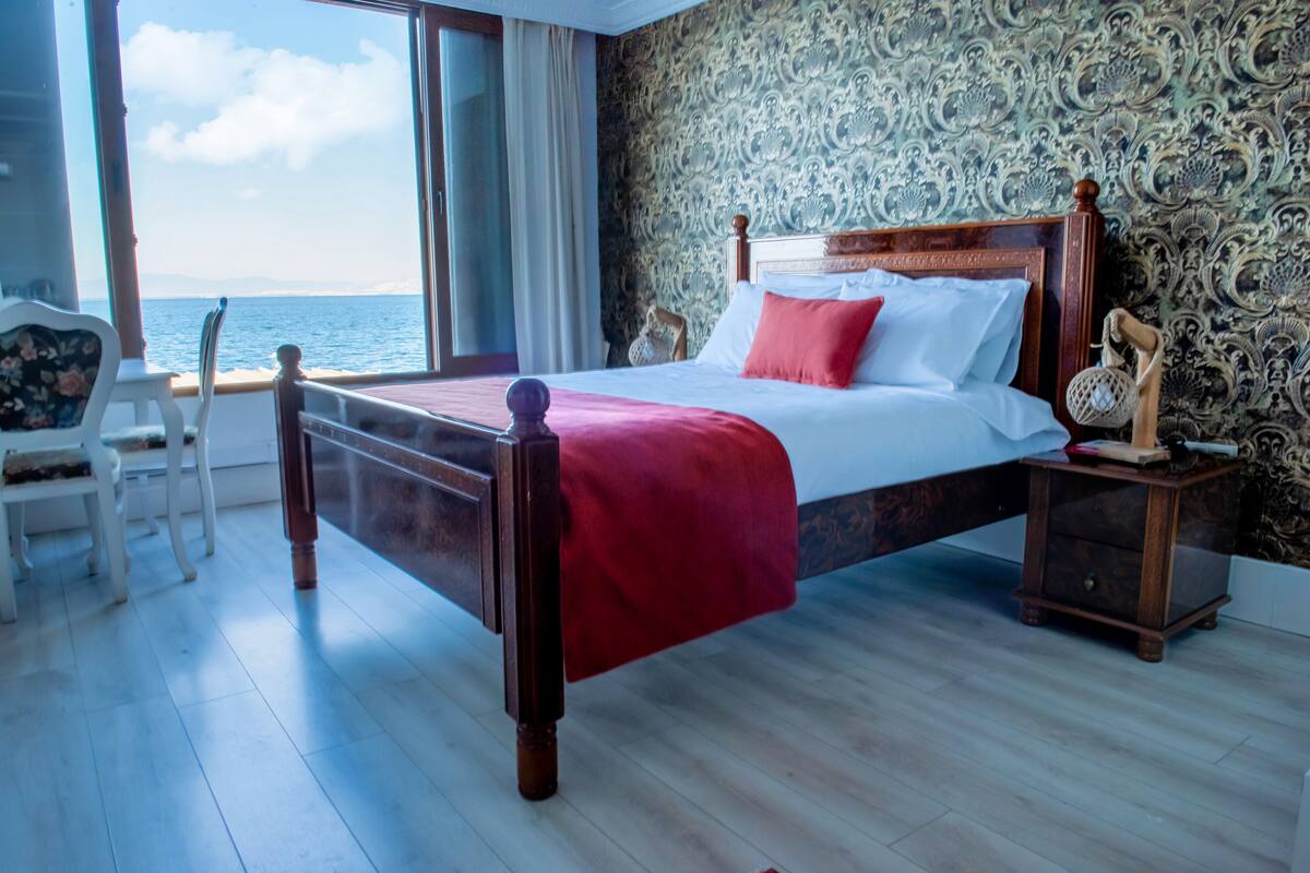 A bedroom with a wooden bed, a nightstand, a chair, and a view of the sea through a large window.