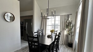 Un coin repas avec une table en bois, des chaises et un lustre. On y trouve également une plante en pot et une fenêtre avec des rideaux.
