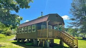 A wooden cabin with a covered porch and a staircase leading up to it, situated on stilts in a grassy area with trees around. - Meadow Riverfront Cabin
 (Nallen)