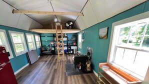 A spacious room with a loft area, wooden flooring, a red refrigerator, and a large window providing natural light. - Meadow Riverfront Cabin
 (Nallen)