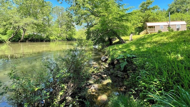 A riverside cabin with a wooden deck and a small porch, surrounded by lush greenery and trees. - Meadow Riverfront Cabin
 (Nallen)