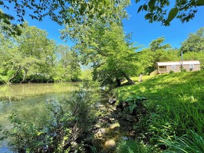 Una cabana a la riba del riu amb terrassa de fusta i petit porxo, envoltada de vegetació abundant i arbres.