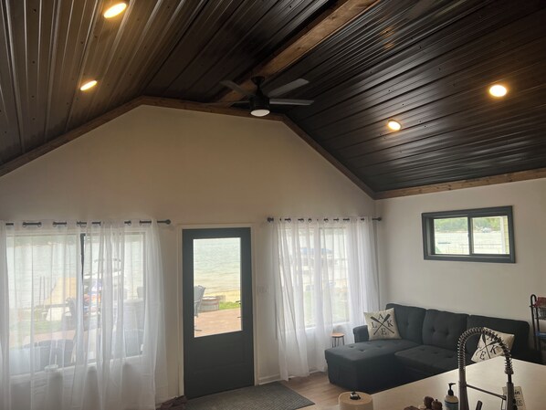 Une pièce chaleureuse avec un plafond en bois sombre, un canapé noir, une petite table et une fenêtre donnant sur l’extérieur.