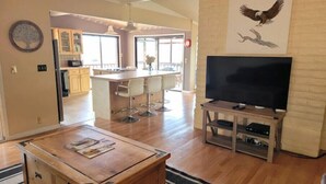 Un vaste espace de vie comprenant un salon avec une table basse en bois, un grand téléviseur à écran plat posé sur un meuble en bois, un coin cuisine avec un bar et un coin repas équipé de tabourets de bar.