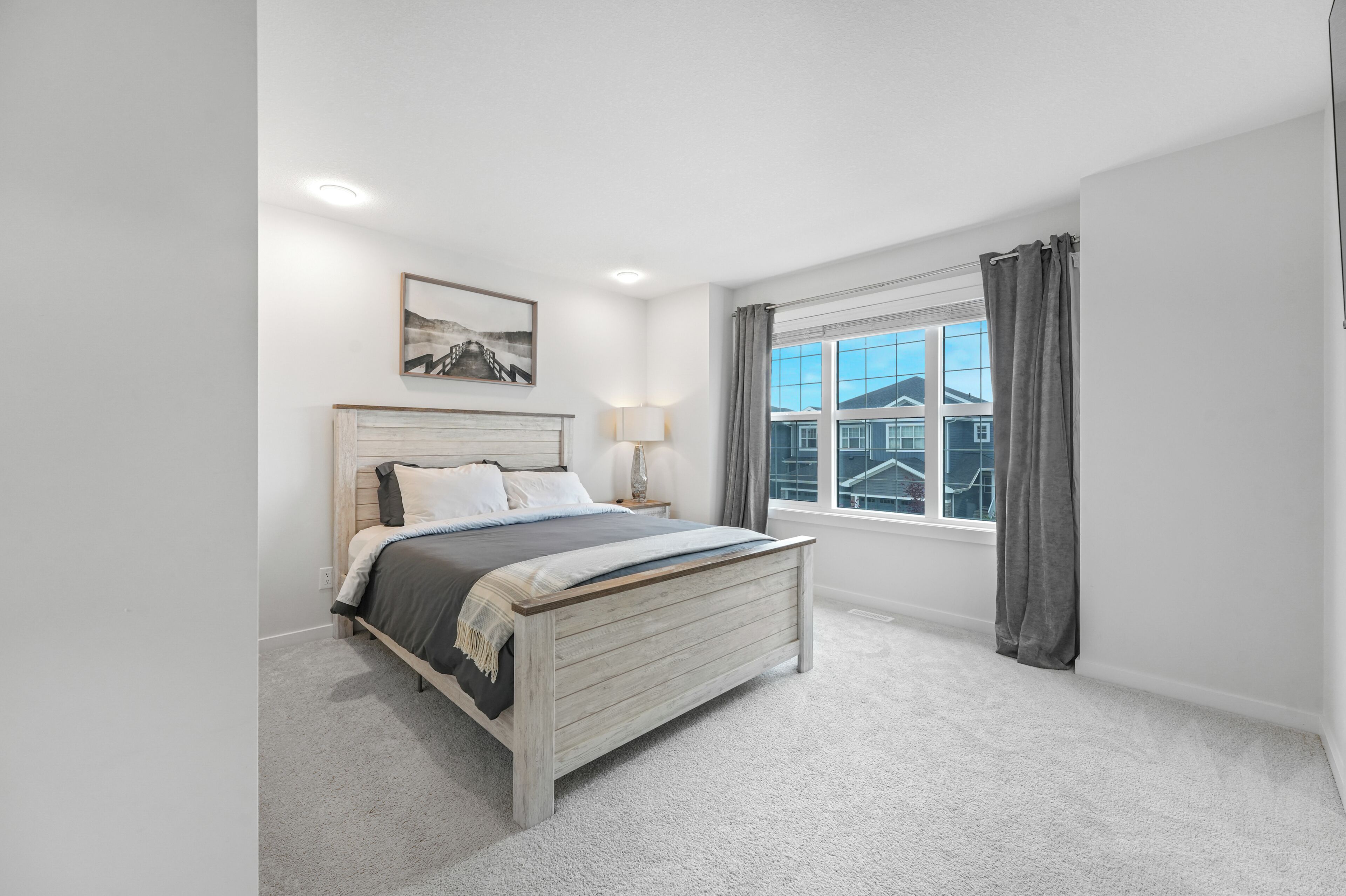 A bedroom with a bed, a window with curtains, a bedside lamp, and a framed picture on the wall.
