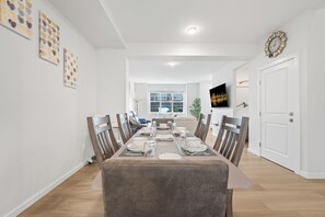 Une salle à manger avec une longue table dressée pour six personnes, entourée de chaises, un tableau représentant un paysage urbain accroché au mur et une horloge au-dessus de la porte.