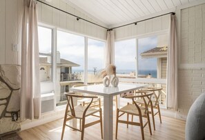A dining area with a white table and four wooden chairs, large windows with curtains, and a view of the sea. - Gorgeous Beachside Bliss Ocean View Walk2Strand (Manhattan Beach)