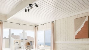 Una sala de estar moderna con un sofá gris, sillones blancos y una zona de comedor con mesa y sillas. Un ventanal ofrece vistas al mar y a un edificio.
