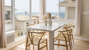 Un comedor con una mesa blanca y sillas de madera, ventanas amplias con cortinas y vistas al mar.