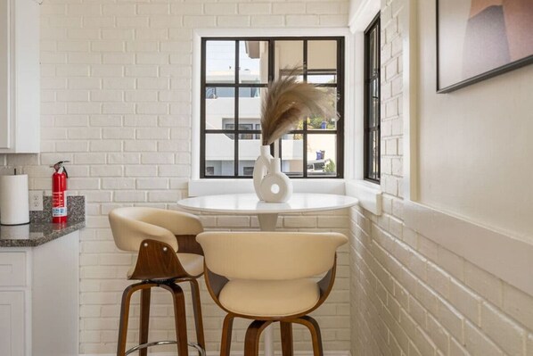 A small kitchen with a white brick wall, a round table with two chairs, a vase with dried plants, and a window with black frames.
